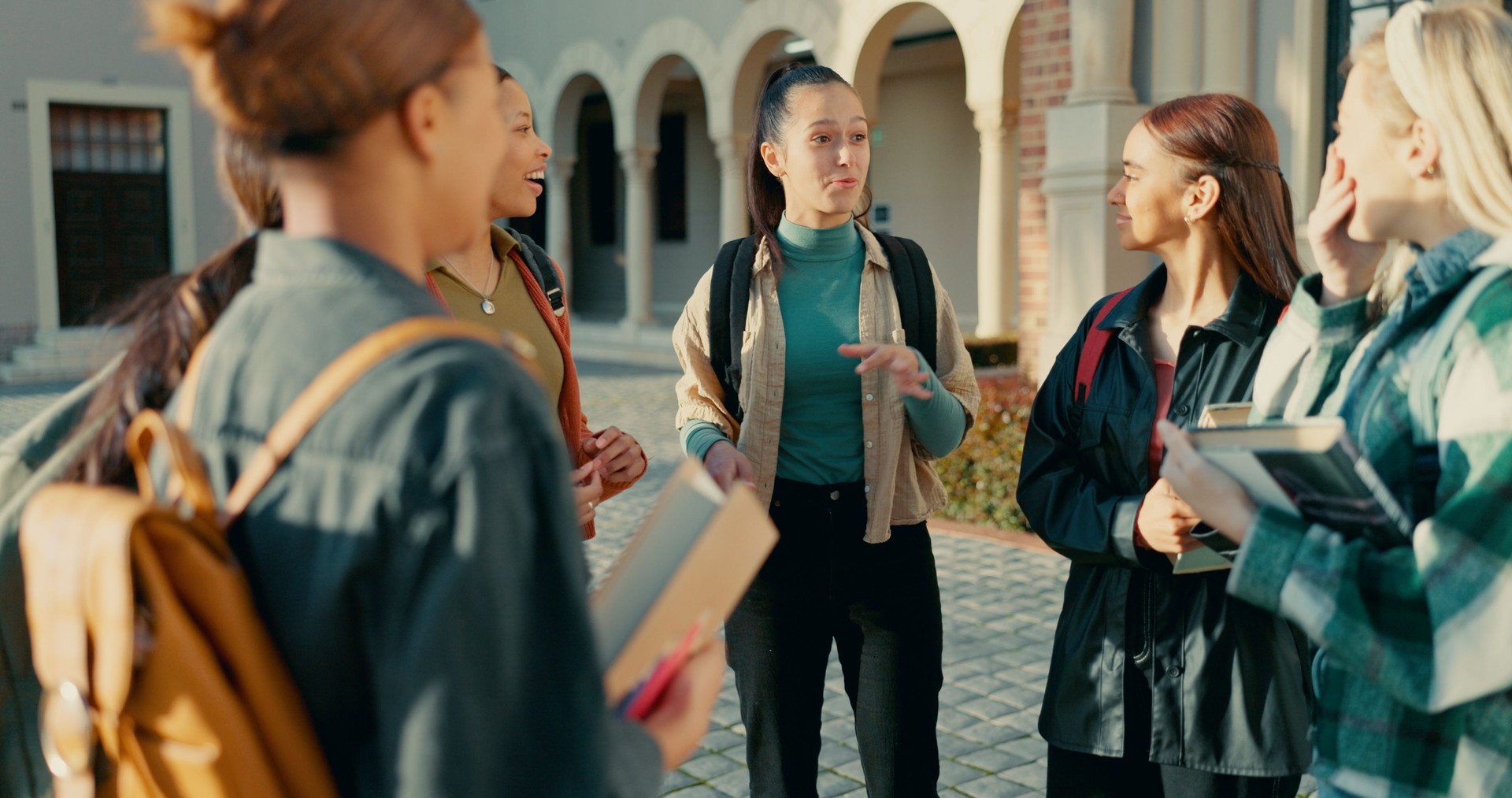 Friends, students and girls at college with talking in circle for funny conversation, university gossip or backpack. Campus, people or diversity with bonding, learning break and happy for scholarship